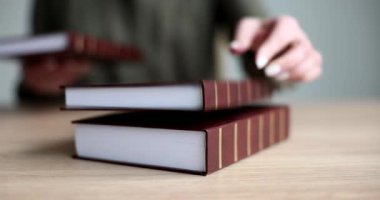 Kütüphaneci, ahşap masaya kahverengi kaplı kalın kitaplar atar. Kadın kütüphanede yavaş çekimde öğrencilere profesyonel malzeme dağıtıyor.