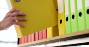 Sekreter sarı klasörde malzeme rafına geri koyar. Arşiv dosyaları yavaş çekimde olan çok renkli halka klasörleri