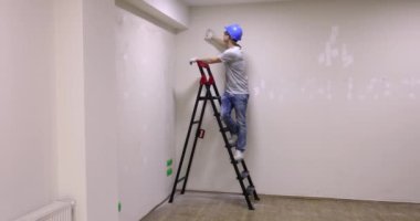 Foreman, tadilat sırasında duvarın sıvanmış halini kontrol etmek için merdivenin üzerinde duruyor. İş kıyafetli adam ağır çekimde gülümseyen jestler gösteriyor.