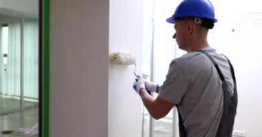 Mavi kasklı, odaklanmış ustabaşı beyaz duvarları silindirle boyuyor. İş kıyafetli bir adam daireyi yavaş çekimde yenilemek için özel bir araç kullanıyor.