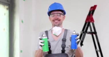 Mutlu ustabaşı yeşil ve mavi şişelerden beyaz boya püskürtür. Koruyucu miğferli ve gözlüklü neşeli adam yavaş çekimde gülümsüyor.