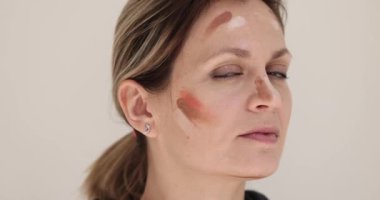 Yüzünde pastel fondöten tonları olan kadın kameraya bakmak için başını çeviriyor. Kadın yüz derisini profesyonel makyaj için yavaş çekime hazırlar.