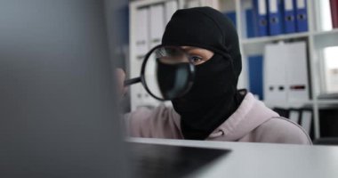 Internet stealing masked person looks at computer screen with magnifying glass. Internet fraud and cybercrime concept