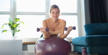 Portrait of smiling woman doing exercise with dumbbells on fitness ball. Fitness equipment for sport activity. Healthy lifestyle concept