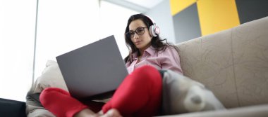 Dizüstü bilgisayar ve kulaklık kullanarak kanepede oturan kadın portresi. Uzaktan çalışma ya da evde boş zaman. Eğlence, uzaklık öğrenme veya çevrimiçi buluşma kavramı