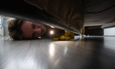 Yatağın altında bir şey aramak için fener kullanan bir kadının yakın çekimi. Koruyucu eldivenli bir kadın yatağın altını temizliyor.