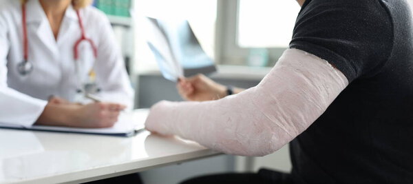 Close-up of man with broken arm at doctor appointment. Doctor at hospital prescribing treatment. Injury, accident, healthcare and medicine concept