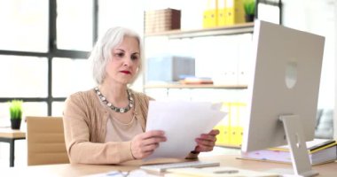 Düşünceli olgun bir kadın iş yerindeki anlaşma belgelerini yavaş çekimde kontrol ediyor. Konsantre çalışanlar masa başında evrak işleriyle uğraşır. Bayan patron iş yerinde.