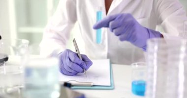 Bilimadamı laboratuvarda incelenen kimyasal reaktörün rengini ve durumunu tanımlıyor. Lastik eldivenli kadın elinde mavi bir sıvıyla ağır çekimde oturuyor.