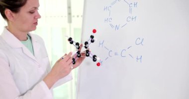 Ciddi kadın, elinde model olan kimyasal bileşik moleküllerini inceliyor. Kimyager, ofiste ağır çekimde beyaz tahtaya yazılmış formüllerden bahsediyor.