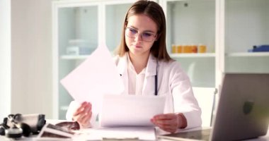 Odaklanmış kadın doktor, açık dizüstü bilgisayarın yanındaki masada oturan hastanın test sonuçlarına bakıyor. Kadın, klinikte ağır çekimde tıbbi evrakları inceliyor.