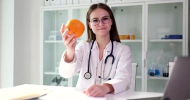 Pozitif kadın doktor olgun turuncuyu elinde tutuyor kameraya bakıyor ve hafifçe gülümsüyor. Steteskoplu neşeli kadın klinikte ağır çekimde oturuyor.