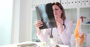 Üniformalı doktor hastayla randevusundan önce eklem röntgenini inceliyor. Kadın klinikteki masada oturuyor ve ağır çekimde radyografa bakıyor.
