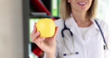 Pozitif kadın doktor klinikte gülümseyen taze elmayı elinde tutuyor. Tıbbi üniformalı neşeli sarışın kadın sağlıklı beslenme öneriyor.