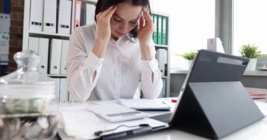 Tired woman with headache at the workplace in office. Depressed tired woman is sitting at table in office. Unhappy overworked young woman at work concept