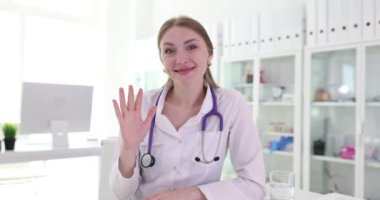 Tıp önlüğü giymiş kadın doktor masada oturmuş gülümsüyor. Tıp bürosundaki neşeli kadın el sallayarak hasta ağır çekimde bekliyor.