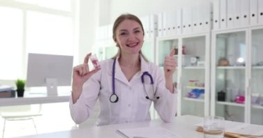 Kadın doktor elinde ilaçla elinde şişeyle baş parmağını kaldırıyor. Neşeli kadın sağlık çalışanı müşterilere yavaş çekimde ilaç reklamı yapıyor.