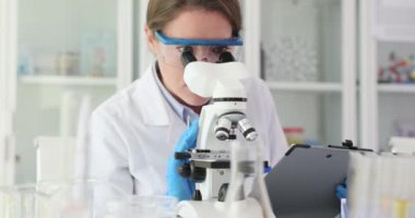 Dişi laboratuvar çalışanı mikroskoba bakıp not alıyor. Tıbbi araştırmacı, detaylı gözlemleri not defterine kaydeden numuneleri inceliyor.