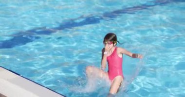 Pembe mayo giymiş küçük bir kız soğuk suya atlıyor. Çocuk havuzun kenarında duruyor ve ferahlatıcı suya atlıyor.