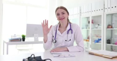 Genç bayan doktor masada oturur ve neşeyle el sallar. Nazik ifadeli kadın doktor hasta yavaş çekimde kameraya el sallıyor.