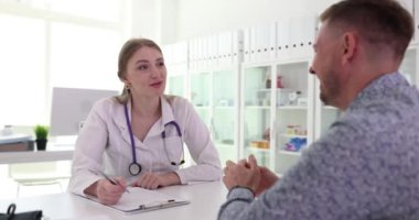 Kadın doktor erkek hasta semptomlarını dikkatle dinler. Profesyonel doktor sohbetin detaylarını yavaş çekimde not defterine kaydeder.