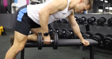 Genç adam, modern spor salonunda yavaş çekimde dambıl sırığı egzersizi yapıyor. Sporcu kulüpte spor yaparken üst vücut geliştirme duruşunu geliştirir