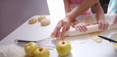 Mom and daughter roll the dough together with a rolling pin on the table, hands close-up, blurry. Teaching a child to cook baked goods