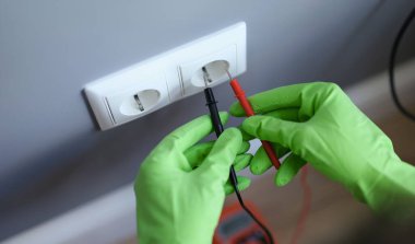 Hands in green gloves plug the voltmeter into an outlet, close-up. Home electrical wiring repair, electrician equipment