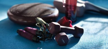 Burgundy skipping rope dumbbells and massager on a blue sports mat, close-up, blurry. Fitness set, sun glare
