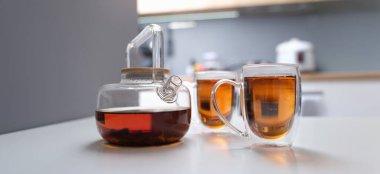 Glass teapot and transparent cups with tea on the table, close-up, blurry. Warming drink at home in the kitchen