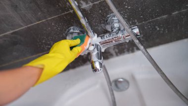 A hand in a yellow glove washes the mixer in the bathroom with a soapy sponge, close-up. Bathroom cleaning, disinfection