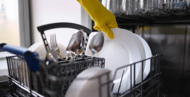A hand in a yellow glove pulls clean dishes out of the dishwasher. Household appliances for home, kitchen utensils
