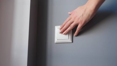 A womans hand presses the switch on the wall, close-up. White light switch on gray wall background, checking electricity