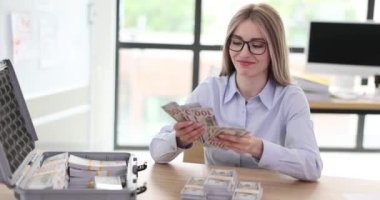 Neşeli iş kadını iş yerinde evrak çantasıyla oturarak para sayıyor. Mutlu kadın banknotlardan hayranı yapıyor zenginliğin tadını çıkarıyor.