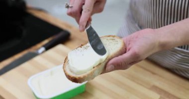Kadın eli, tezgahta bıçakla yumuşacık tereyağı yayarak ekmeği tutuyor. Önlüklü ev hanımı öğle yemeği için lezzetli sandviçler hazırlar.