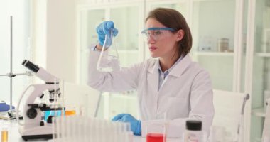 Kadın laboratuvar çalışanı iş yerinde şeffaf sıvı reaksiyonlu matara tutuyor. Dikkatli bir kadın laboratuarda yapılan deneylerden sonra değişiklikleri inceler.