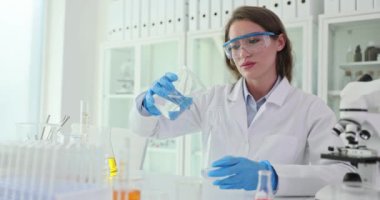 Laboratuvar çalışanı deney şişesinden cam bardağa sıvı doldurup araştırma için hazırlanıyor. Üniformalı özenli bir kadın iş yerinde oturarak deney yapıyor.