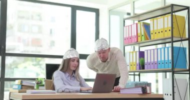 Alüminyum folyo şapkalı ofis çalışanları iş yerindeki laptoptaki verileri kontrol ediyor. Kepçeli bir adam iş sırasında bayan meslektaşının güvenliğinden endişe ediyor.