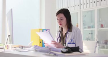 Steteskoplu kadın doktor klinikteki iş yerinde oturan kağıtları inceliyor. Üniformalı gevşek saçlı kadın doktor yorgunluk ve baş ağrısı çekiyor.