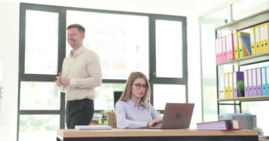 İşadamı modern şirket ofisinde dizüstü bilgisayarda yazan kadın asistanla konuşuyor. Dost canlısı erkek patron, meşgul kadın çalışana iş gücü veriyor.