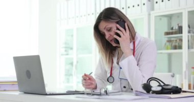 Kadın doktor telefonda konuşuyor. Hastanede işyerinde oturuyor. Steteskopla ilgili kadın doktor hastaya çevrimiçi danışmanlık hizmeti sunuyor.