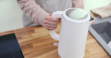 Genç bir kadın evdeki mutfağa elektrikli su ısıtıcısı yerleştiriyor. Ev hanımı fişe takmaya hazır ve taze demlenmiş çayın sıcaklığını tahmin ederek suyu ısıtmaya hazır.