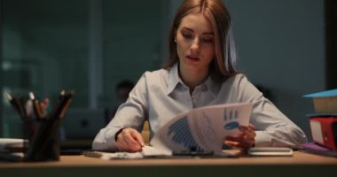 Yorgun genç bayan işçi raporları çizelgelerle inceliyor. Bayan, gece geç saatlerde ofiste evrak işleriyle uğraşırken akıllı telefona bakıyor.