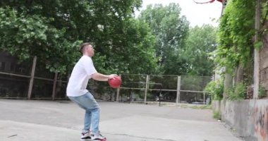 Atletizm trenleri ağır basketbol topunu yavaş çekimde potaya fırlatıyor. Sporcu yaz günü açık hava sahasında aktif antrenman yapıyor.