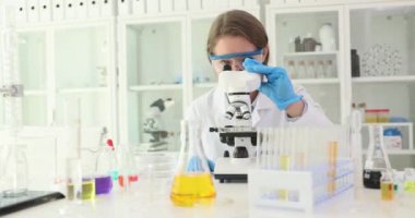 Kadın araştırmacı örnekleri mikroskop kullanarak analiz ediyor. Kadın bilim adamı, meslektaşının laboratuvara ağır çekim girdiğini onaylamak için el sallıyor.