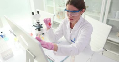 Kadın kimyagerin elinde kırmızı reaktörle dolu bir kap var. Bilim adamı, reaksiyonun kimyasal özellikleri hakkında ayrıntılı bilgi bulmak için bilgisayarı kullanır