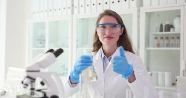 Kadın bilim adamı laboratuarda elinde bir şişe sıvı tutarken parmaklarını gösterdi. Koruyucu üniformalı kadın teknisyen sıvılarla deney yapıyor.