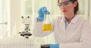 Kadın bilim adamı laboratuardaki konik şişede sarı sıvı maddeyi sallıyor. Kadın kimyager laboratuvarda çalışan sıvı test süresini kontrol ediyor.