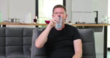 Susamış genç adam şeffaf şişeden su içiyor ve tatmin oluyor. Oturma odasındaki kanepede oturan adam sıvı dengesini koruyor.