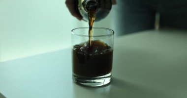 Elleri şeffaf bir şişe tutuyor ve masaya tatlı bir içecek koyuyor. Koyu renkli ve lezzetli bir içecek sunar.